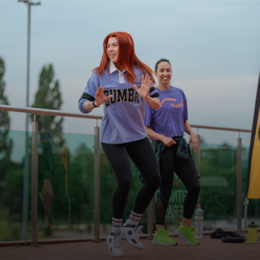 Dvi sportinę aprangą vilkinčios moterys veda zumbos pamoką lauke ant asfaltuotos dangos. Viena jų entuziastingai šypsosi šokdama. Fone matyti geltona vėliava ir medžiai - tai rodo, kad vyksta grupinis fitneso renginys.
