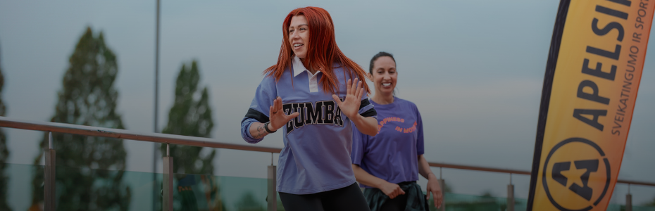 Dvi moterys, viena iš jų raudonplaukė, vilkinti violetinius marškinėlius su užrašu "ZUMBA", šypsosi ir šoka lauke šalia sveikatingumo plakato "APELSINAS", o fone matyti medžiai ir debesuotas dangus.
