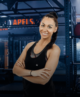 Moteris ilgais tamsiais plaukais, supintais į kasą, vilkinti juodą liemenėlę, šypsosi sukryžiavusi rankas sporto salėje. Fone matyti sporto salės įranga ir šviečiantis užrašas "APELSIN".