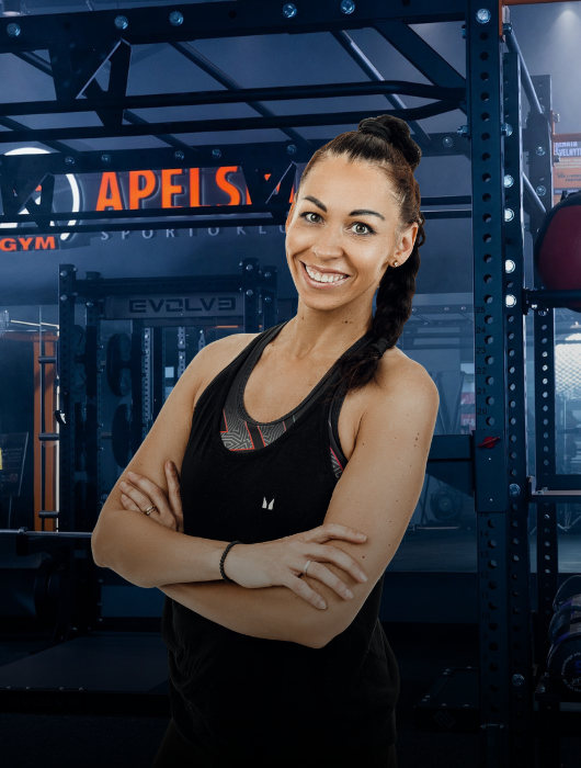 Moteris ilgais tamsiais plaukais, supintais į kasą, vilkinti juodą liemenėlę, šypsosi sukryžiavusi rankas sporto salėje. Fone matyti sporto salės įranga ir šviečiantis užrašas "APELSIN".