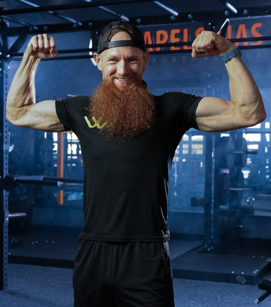 Žmogus su ilga raudona barzda ir juoda kepuraite sulenkia bicepsą šypsodamasis sporto salėje. Jie dėvi juodus marškinėlius ir šortus, juos supa sporto salės įranga ir mėlynas aplinkos apšvietimas.