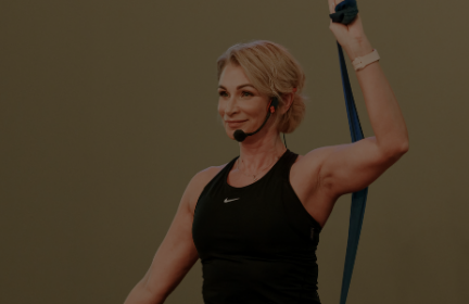 Sportine apranga pasipuošusi moteris šypsosi virš galvos laikydama pasipriešinimo juostą. Ji nešioja ausinių mikrofoną, o tai rodo, kad ji gali vesti kūno rengybos pamoką. Fone yra neutrali patalpų aplinka.