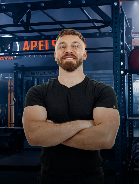 Šypsodamasis trumpai kirptų plaukų ir barzdotas vyras, sukryžiavęs rankas, užtikrintai stovi menkai apšviestoje sporto salėje, apsuptas sunkiosios atletikos įrangos ir stelažų.