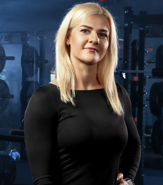 Moteris ilgais šviesiais plaukais, vilkinti juodus marškinius ilgomis rankovėmis, užtikrintai stovi sporto salėje. Fone yra svarmenys ir treniruokliai.