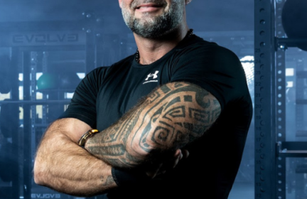 Raumeningas vyras nuskusta galva ir tatuiruotomis rankomis užtikrintai stovi sporto salėje ir šypsosi sukryžiavęs rankas. Jis vilki juodus marškinėlius, o neryškiame fone matosi treniruokliai.