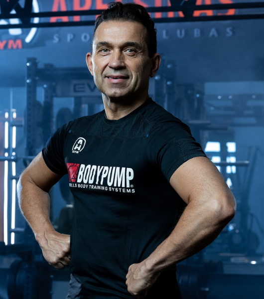 Asmuo sporto salėje, vilkintis juodus „BodyPump“ marškinėlius, pozuoja užtikrintai laikydamas rankas ant klubų. Matosi treniruokliai ir iškaba su užrašu „GYM“ fone.