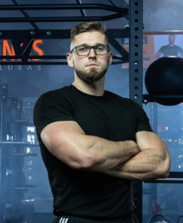 Vyras su akiniais ir barzda užtikrintai stovi sporto salėje, vilkėdamas juodus marškinius ir juodas kelnes. Jam sukryžiuotos rankos, o fone matosi sporto salės įranga.