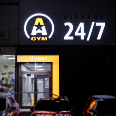 Įėjimas į sporto salę su ryškiu užrašu „24/7“ ir „A Gym“ ant pastato fasado. Įėjimas apšviestas, o pirmame plane matosi keli stovintys automobiliai.