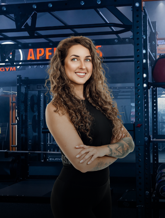 Moteris ilgais garbanotais plaukais ir tatuiruotėmis ant dilbio šypsodamasi stovi sukryžiavusi rankas sporto salėje, apsupta sunkiosios atletikos įrangos ir stovų.