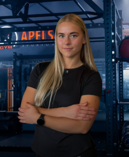 Moteris ilgais šviesiais plaukais užtikrintai stovi sudėjusias rankas sporto salėje. Ji vilki juodus marškinėlius ir išmanųjį laikrodį. Sporto salėje yra treniruoklių ir tamsiai mėlynos bei oranžinės spalvos.