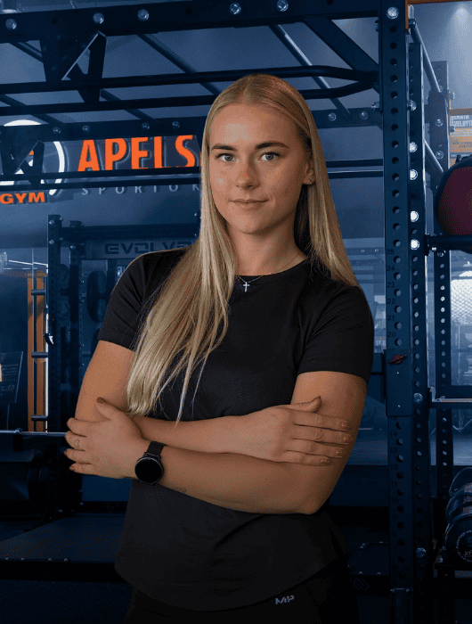 Moteris ilgais šviesiais plaukais užtikrintai stovi sudėjusias rankas sporto salėje. Ji vilki juodus marškinėlius ir išmanųjį laikrodį. Sporto salėje yra treniruoklių ir tamsiai mėlynos bei oranžinės spalvos.