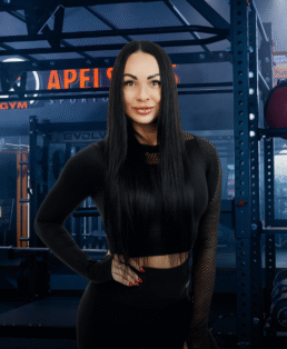 Moteris ilgais juodais plaukais, vilkinti juodą sportinę aprangą, užtikrintai stovi sporto salėje, kurios fone matoma sunkiosios atletikos įranga ir stendai.