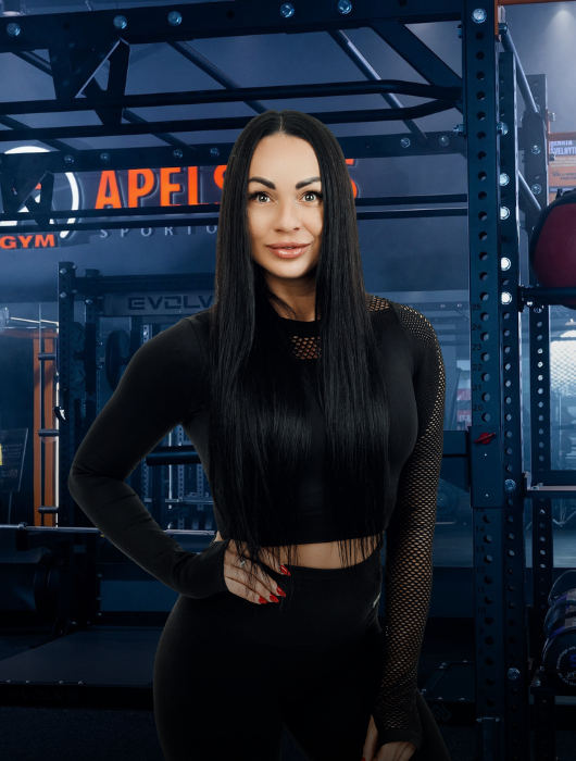 Moteris ilgais juodais plaukais, vilkinti juodą sportinę aprangą, užtikrintai stovi sporto salėje, kurios fone matoma sunkiosios atletikos įranga ir stendai.