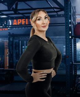 Juodais sportiniais drabužiais apsirengusi moteris užtikrintai stovi rankomis ant klubų sporto salėje, kurios fone matoma sunkiosios atletikos įranga ir užrašas "APEX".