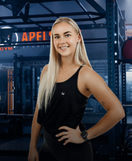 Besišypsanti jauna moteris ilgais šviesiais plaukais užtikrintai stovi sporto salėje, vilkėdama juodą liemenėlę ir juodas tampres, ranką laikydama ant klubo. Fone matyti sporto salės įranga ir svarmenys.