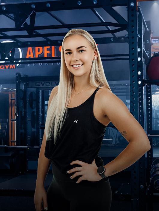 Besišypsanti jauna moteris ilgais šviesiais plaukais užtikrintai stovi sporto salėje, vilkėdama juodą liemenėlę ir juodas tampres, ranką laikydama ant klubo. Fone matyti sporto salės įranga ir svarmenys.