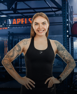 Besišypsanti moteris ilgais plaukais ir tatuiruotėmis ant rankovių užtikrintai stovi sporto salėje, vilkėdama juodą sportinę liemenėlę ir išmanųjį laikrodį, o fone matoma sporto salės įranga.