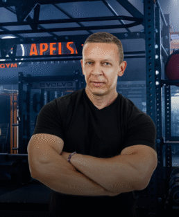 Raumeningas vyras juodais marškinėliais stovi sukryžiavęs rankas sporto salėje, kurios fone matyti treniruokliai ir užrašas "APELS".