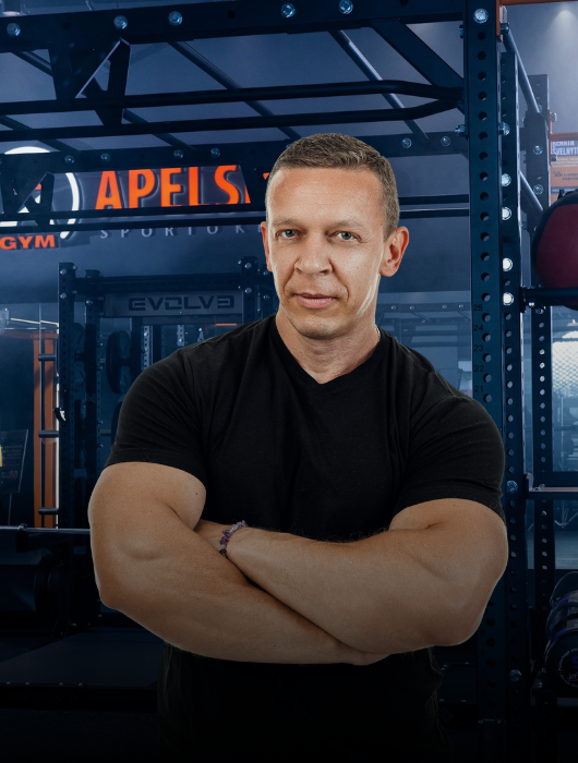 Raumeningas vyras juodais marškinėliais stovi sukryžiavęs rankas sporto salėje, kurios fone matyti treniruokliai ir užrašas "APELS".