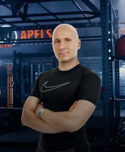 Pliktelėjęs vyras su juodais "Nike" marškinėliais stovi sukryžiavęs rankas sporto salėje, kurios fone matomi svarmenų stovai ir treniruokliai. Apšvietimas vėsus, už jo matomas sporto salės užrašas "APELSIN".