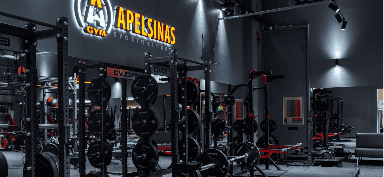 Modernus sporto salės interjeras su įvairia sunkiosios atletikos įranga, įskaitant suolus, lentynas ir svarmenis. Sienos tamsios su ryškiu apšvietimu, o iškaboje šviečiančiomis raidėmis parašyta „Apelsinas Gym“.