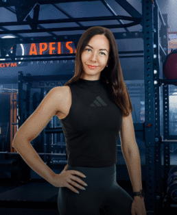 Ilgais rudais plaukais moteris, vilkinti juodą palaidinę be rankovių ir tamsius kedus, užtikrintai stovi sporto salėje. Ji šypsosi į kamerą, vieną ranką laikydama ant klubo, fone matomi sporto salės įrenginiai.