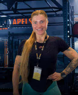 Besišypsanti moteris ilgais šviesiais plaukais, surištais į kuodą, stovi sporto salėje, dėvi dirželį su vardiniu ženkleliu, juodus marškinėlius, arbatžoles ir išmanųjį laikrodį. Fone matyti sporto salės įranga ir stelažai.