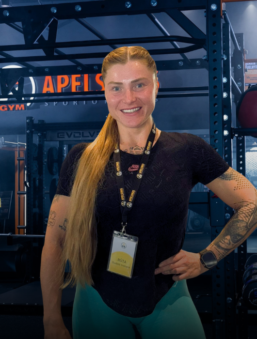 Besišypsanti moteris ilgais šviesiais plaukais, surištais į kuodą, stovi sporto salėje, dėvi dirželį su vardiniu ženkleliu, juodus marškinėlius, arbatžoles ir išmanųjį laikrodį. Fone matyti sporto salės įranga ir stelažai.