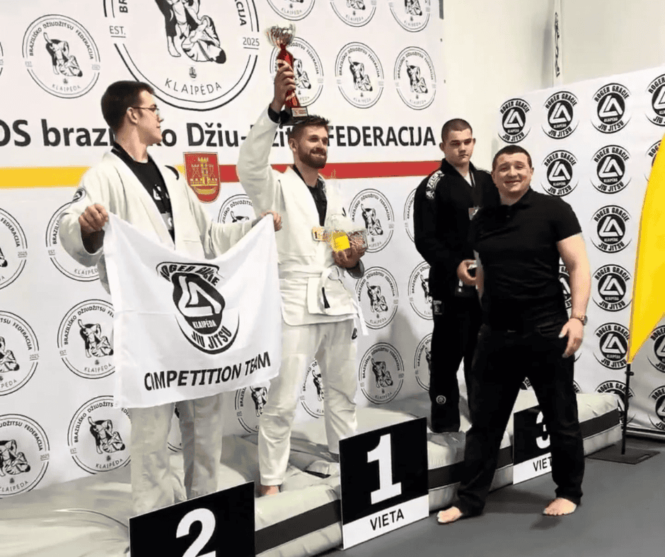Keturi vyrai stovi ant kovos menų pakylos. Pirmoje vietoje esantis vyras, vilkintis baltą kostiumą, laiko trofėjų ir medalį. Antros vietos laimėtojas laiko komandos vėliavą, o trečios vietos laimėtojas stovi šalia jų. Besišypsantis vyras yra priešais pakylą.