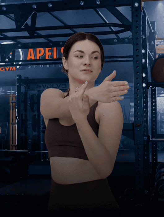 Ruda sportine apranga vilkinti moteris ištiesia ranką ant krūtinės sporto salėje, apsuptoje treniruoklių ir užrašo "APELIS" fone.