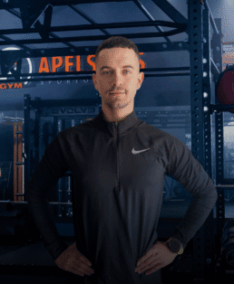 Vyras, vilkintis juodą sportinę "Nike" striukę, rankas laikydamas ant klubų užtikrintai stovi sporto salėje, kurios fone matomi svarmenų stovai ir įranga.