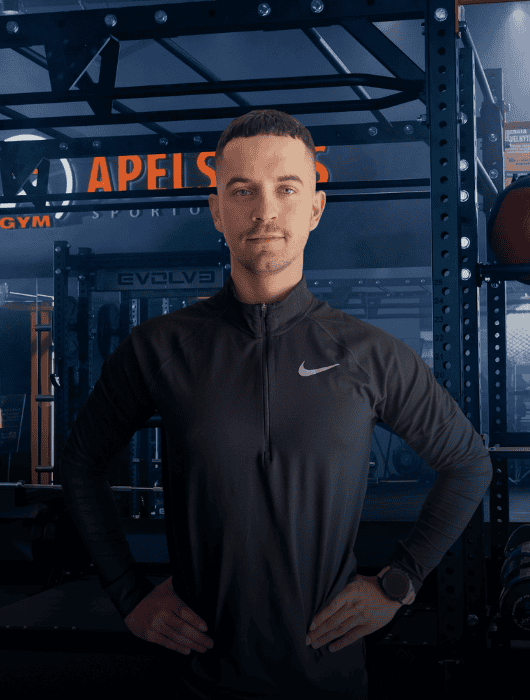 Vyras, vilkintis juodą sportinę "Nike" striukę, rankas laikydamas ant klubų užtikrintai stovi sporto salėje, kurios fone matomi svarmenų stovai ir įranga.