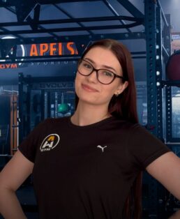 Moteris ilgais rudais plaukais ir akiniais, vilkinti juodus marškinėlius su sporto salės logotipu, užtikrintai stovi sporto salėje, apsupta treniruoklių ir fone esančios metalinės stovo konstrukcijos.