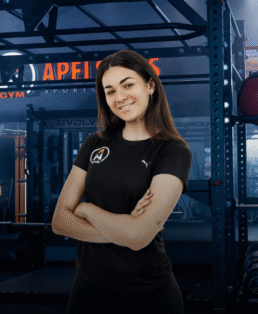 Moteris ilgais tamsiais plaukais pasitikinčiai stovi sukryžiavusi rankas ir šypsosi sporto salėje. Ji vilki juodus marškinėlius su logotipais, o fone matyti sporto salės įranga.