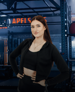 Moteris ilgais rudais plaukais, vilkinti juodą sportinę aprangą ir su grandinėle ant kaklo, pasitikinčiai stovi rankomis ant klubų modernioje sporto salėje, kurios fone matyti treniruokliai.