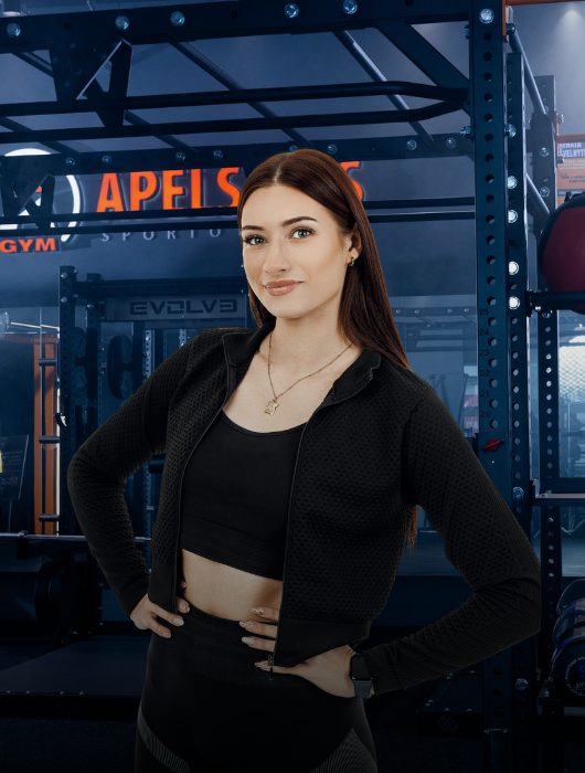 Moteris ilgais rudais plaukais, vilkinti juodą sportinę aprangą ir su grandinėle ant kaklo, pasitikinčiai stovi rankomis ant klubų modernioje sporto salėje, kurios fone matyti treniruokliai.