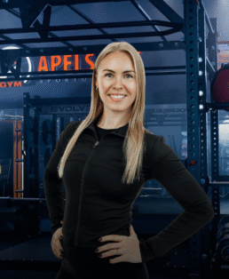 Besišypsanti moteris ilgais šviesiais plaukais, vilkinti juodą sportinę liemenėlę su užtrauktuku, pasitikinčiai stovi rankomis ant klubų modernioje sporto salėje su treniruokliais fone.