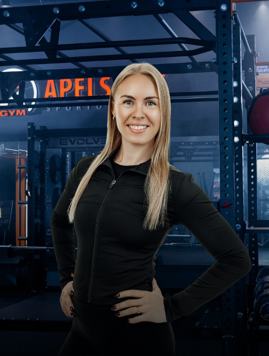 Besišypsanti moteris ilgais šviesiais plaukais, vilkinti juodą sportinę liemenėlę su užtrauktuku, pasitikinčiai stovi rankomis ant klubų modernioje sporto salėje su treniruokliais fone.