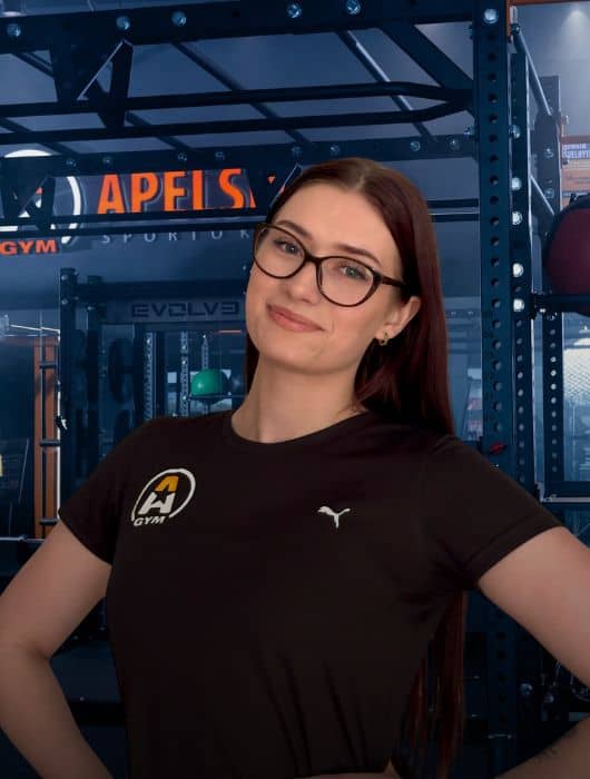 Moteris ilgais rudais plaukais ir akiniais, vilkinti juodus marškinėlius su sporto salės logotipu, užtikrintai stovi sporto salėje, apsupta treniruoklių ir fone esančios metalinės stovo konstrukcijos.
