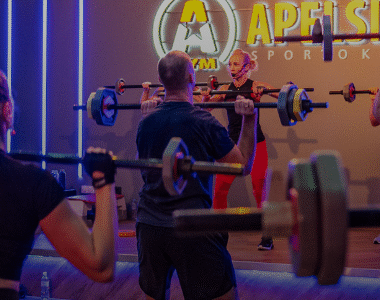 Grupinis fitneso užsiėmimas sporto salėje, kur žmonės kelia štangas virš pečių, stovi priešais instruktorių ant scenos po neoninėmis šviesomis ir dideliu užrašu "Apelsinas Sporto Klubas".