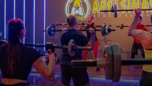 Grupinis fitneso užsiėmimas sporto salėje, kur žmonės kelia štangas virš pečių, stovi priešais instruktorių ant scenos po neoninėmis šviesomis ir dideliu užrašu "Apelsinas Sporto Klubas".