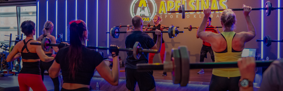 Grupinis fitneso užsiėmimas sporto salėje, kur žmonės kelia štangas virš pečių, stovi priešais instruktorių ant scenos po neoninėmis šviesomis ir dideliu užrašu "Apelsinas Sporto Klubas".