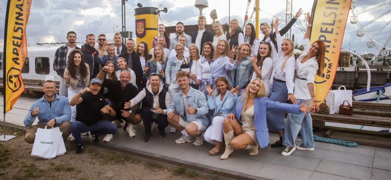Didelė grupė besišypsančių žmonių pozuoja lauke prie prisišvartavusios valties, laikydami taikos ženklus ir mojuodami rankomis. Fone matyti geltonos vėliavos su "Adelsinas" ženklu ir debesuotas dangus.