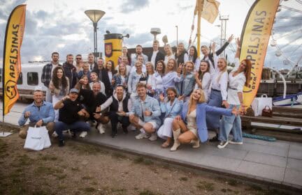 Didelė grupė besišypsančių žmonių pozuoja lauke prie prisišvartavusios valties, laikydami taikos ženklus ir mojuodami rankomis. Fone matyti geltonos vėliavos su "Adelsinas" ženklu ir debesuotas dangus.