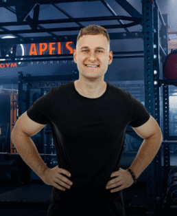 Besišypsantis vyras su juodais marškinėliais stovi su rankomis ant klubų sporto salėje, kurios fone matomi svarmenys, metaliniai stelažai ir treniruokliai.