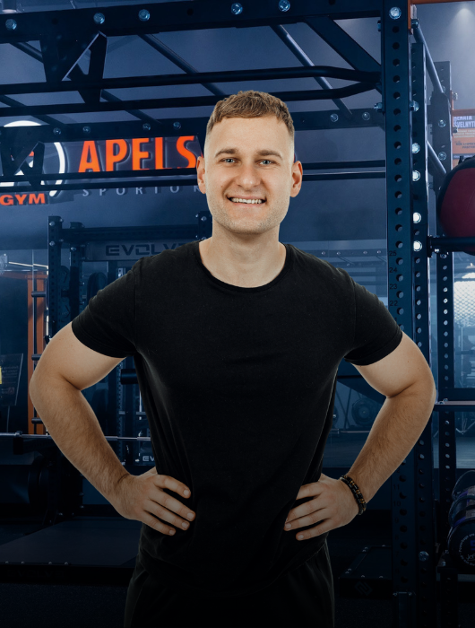 Besišypsantis vyras juodais marškinėliais stovi rankomis ant klubų modernioje sporto salėje, apsuptas sporto įrangos ir fone matomo užrašo "APELS".