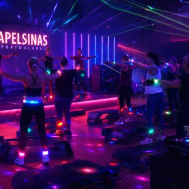 Tamsioje sporto salėje, apšviestoje spalvotomis neoninėmis šviesomis ir lazerių spinduliais, vyksta grupiniai fitneso užsiėmimai, o kampe groja didžėjus. Scenoje šviečiančiomis raidėmis matyti logotipas "APELSINAS GYM". Dalyviai dėvi šviečiančius aksesuarus.