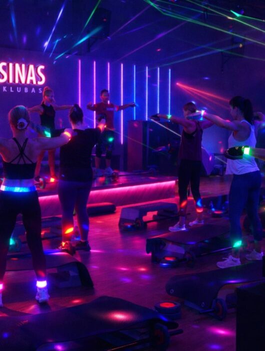 Tamsioje sporto salėje, apšviestoje spalvotomis neoninėmis šviesomis ir lazerių spinduliais, vyksta grupiniai fitneso užsiėmimai, o kampe groja didžėjus. Scenoje šviečiančiomis raidėmis matyti logotipas "APELSINAS GYM". Dalyviai dėvi šviečiančius aksesuarus.