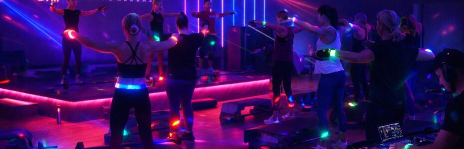 Tamsioje sporto salėje, apšviestoje spalvotomis neoninėmis šviesomis ir lazerių spinduliais, vyksta grupiniai fitneso užsiėmimai, o kampe groja didžėjus. Scenoje šviečiančiomis raidėmis matyti logotipas "APELSINAS GYM". Dalyviai dėvi šviečiančius aksesuarus.